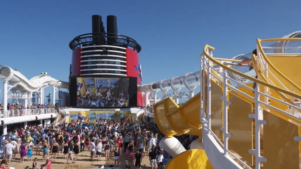 Vue depuis le pont 11 de la piscine avec des passagers jouant dans la piscine et se relaxant sur des bains de soleil.  Vue sur le toboggan aquatique AquaDuck et l'entonnoir emblématique de Disney Cruise Line